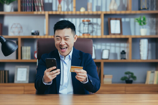 Young Handsome Asian Man In Suit In The Office At Work Holding A Credit Card And Shopping, Ordering Online From The Phone, Happy