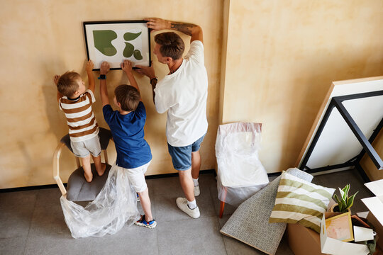 Top view portrait of father with two sons hanging picture on wall together while moving to new home, copy space