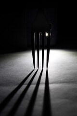 Tenedor para manipular comidas en contraluz entre sus dientes forma una figura en sombra reflejada sobre el piso, presenta un hermoso diseño abstracto con fondo negro - obrazy, fototapety, plakaty