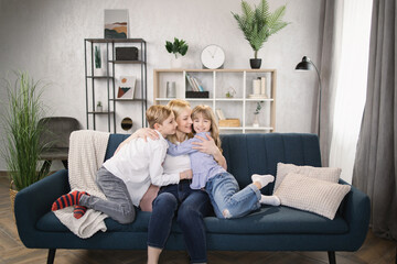 Happy little kids sit on couch hug cuddle excited young mom or nanny show love and care, overjoyed small children relax with mother embrace share sweet tender family moment in living room at home