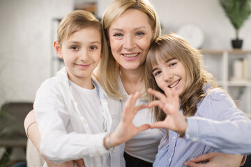Obraz na płótnie Canvas Cute little daughter with son and mother join hands in shape of heart as concept of mom and kids love care support, smiling mum and her kids looking at camera posing together for headshot portrait