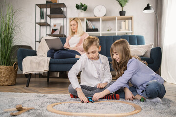 Cute little preschooler boy and girl siblings sit on warm floor play at home together, happy mother relax on sofa on background, smiling small brother and sister involved in activity in living room