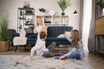 Cheerful little siblings sitting on warm heated wooden floor, chatting communicating playing with a wooden toy train while happy young mom shopping with digital tablet. sitting on sofa and waving hand