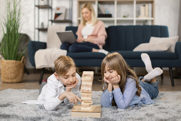 Little children sit on warm floor in living room play construct with wooden building blocks bricks together make a tower, young mom rest on sofa in background, Caucasian family relax at home