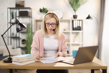 Smiling Caucasian woman writing notes while using laptop computer at bright office or home interior. Pretty female software developer using modern laptop for remote work at home.