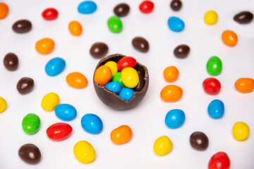 Half of a cracked chocolate egg filled with multi-colored candies on a white background with multi-colored candies, top view. Treats for children. Sweet dessert for kids. Sweet Easter tradition.