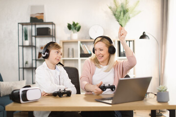 Mother with son sitting together at the table and playing video games with joysticks. Mom raised her hands up and screaming while winning the game. Woman helps to play her son.