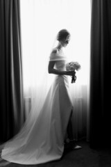 A bride in a white dress stands near the window and looks at the bouquet