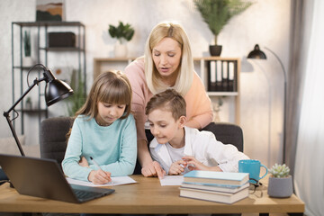 Fototapeta premium Mother teach young son and daughter homework, using digital laptop at home. E-learning lifestyle, happy family homeschooling, children remote distance education, or internet technology concept