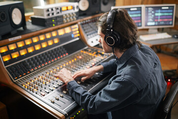 Portrait of young man wearing headphones and operating buttons and toggles at digital audio...