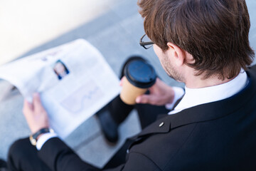 Portrait of a handsome stylish businessman reading information about finance news in newspaper outdoors with coffee in front of  building city background. network technology, internet, successful man.