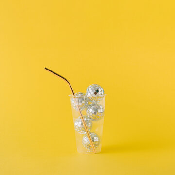 Transparent plastic drinking cup filled with shiny disco balls and golden straw on yellow background.