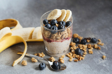 granola with banana and blueberries with yogurt in glass cup on gray background, healthy diet breakfast muesli with yogurt and fruits