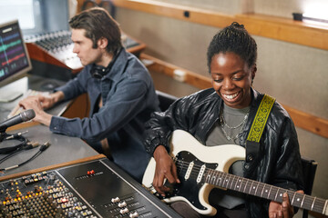 Portrait of smiling black woman playing guitar while producing music in professional recording...