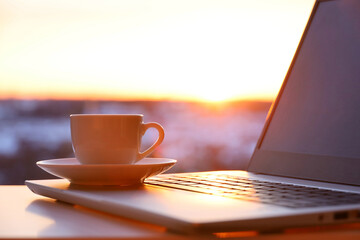 Coffee or tea cup on laptop against the window in sunshine. Cozy workplace in home office, inspiring view to morning city