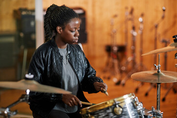 Side view portrait of black young woman playing drums in recording studio and making rock music,...
