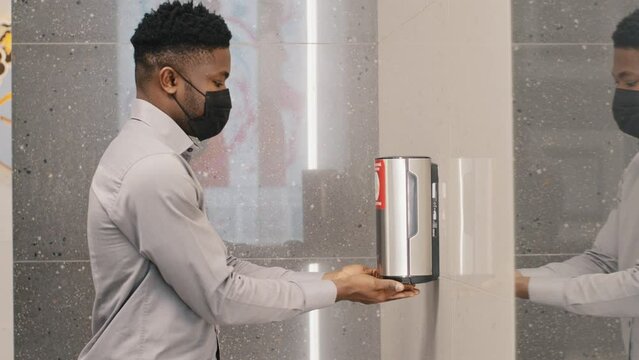 Young african american man in protective mask stands in public toilet dries hands under jet of hot air using hand dryer surprised by modern technology touchless washing hygiene and sanitation concept