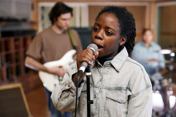 Waist up portrait of young black woman singing to microphone while recording music with band in professional studio