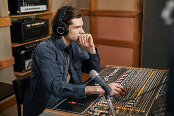 Side view portrait of focused young man wearing headphones at audio workstation in professional recording studio, music production and artistry