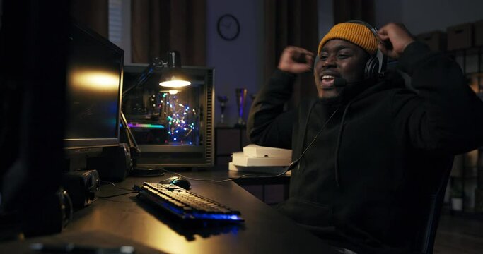 Smiling man in yellow cap with headset pushes back on chair from computer desk, hands high in the air, beats his chest with joy, game won, high salary, dark room lit by purple led lights.