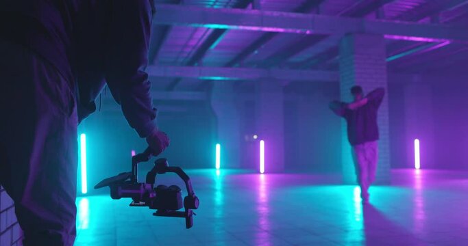 Backstage of video shooting. Cameraman filming hip-hop guy dancer at underground garage space with neon light lamps. Operator moves with image stabilizer in hands