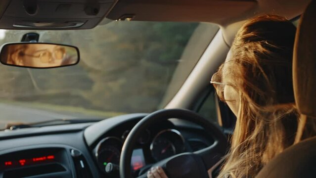 Rear view of happy young blonde in stylish sunglasses dancing to the music while driving her car at sunset. Pretty woman listening to enegetic music and smiling. Concept of joy and happiness