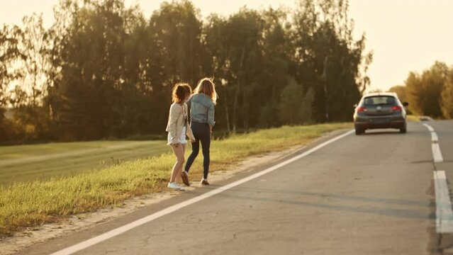 Young sexy girls dressed in casual summer clothes hitchhiking on the road raising their thumbs up. Pretty women travelers waiting for car s at roadside. Concept of freedom and travelling.