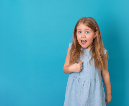 Little surprised girl in blue dress with eyes and mouth wide open asks who me pointing at herself with copy space on blue background.