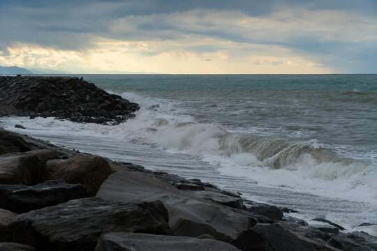 Storm waves roll over large rocks on the seashore with dark clouds and a clear sun in the sky