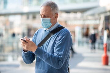 Covid coronavirus concept, masked elder man using his smartphone outdoor