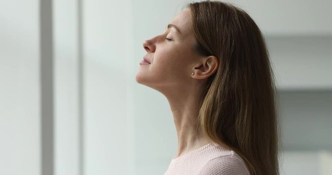 Calm young woman taking deep breath of fresh air meditating with eyes closed standing indoor, close up side profile view. Enjoy moment of peace, do mental relaxation exercises, feels no stress concept