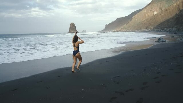 Young woman in swimsuit runs along the black volcanic black sand beach Benijo in the north of Canary island Tenerife