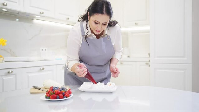 a female chef adds jam from a pastry bag to Anna Pavlova cakes in the kitchen.
