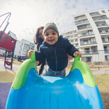 Happy African American Little Boy With A Grey Hat In Front Of His Mother Climbing On A Blue Slide In The Park Buildings Background Cowboy Shot. High Quality Photo