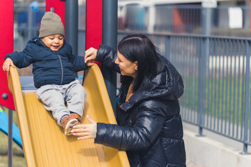 dark-haired woman with a long black coat looking at a sweet little boy who is sliding down on a...