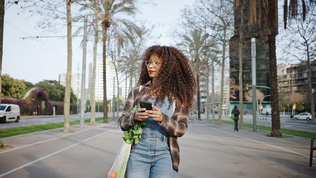 A girl wearing denim overalls and glasses walks along streets of barcelona with a phone in her hands. Happy youg tourist