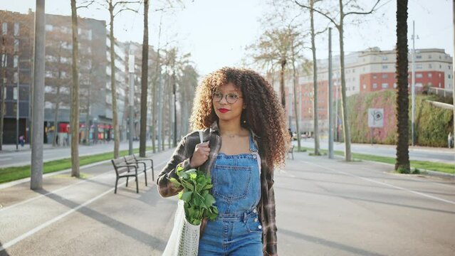 Young long-haired mixer race girl in glasses and denim overalls walks through the city with a shopping bag with fresh greens on her shoulder. Vegetarian and tourism concept