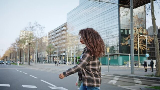 Modern confident woman crosses the city street on the way from the market with a bag of greens on her shoulder. Long-haired curly woman wears glasses and denim overalls, walking in the summer city