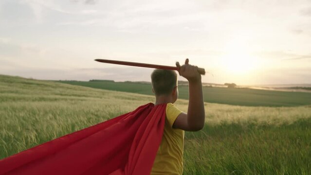 Child boy plays superhero. Child Game. Boy in red cloak stands with sword raised in his hand up sword, on field, depicting medieval knight. Child waves toy sword, childhood dreams. Imagination