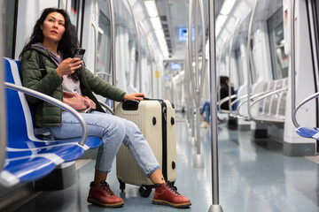 Asian woman with suitcase sitting in subway car and using her smartphone