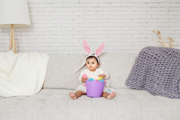 Cute Indian baby girl with pink bunny ears and basket of colorful eggs celebrating Easter holiday.