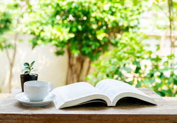Cup of coffee and opened book on wooden table in front yard.
