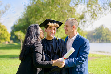 Young recently graduated boy, dressed in cap and gown, with his degree in hands, celebrating with his multi ethnic  family on the university campus. Very happy expression, achievement