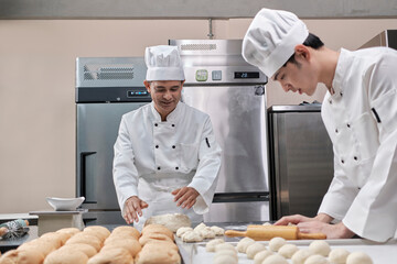 Two professional Asian male chefs in white cook uniforms and aprons are kneading pastry dough and eggs, preparing bread and fresh bakery food, baking in oven at stainless steel kitchen of restaurant.