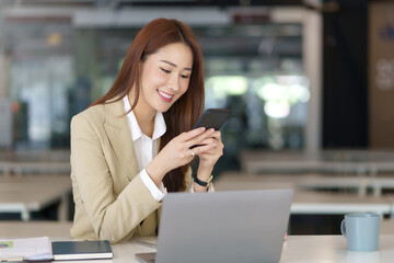 Charming young Asian business woman with a smile sitting using smartphone at the office.