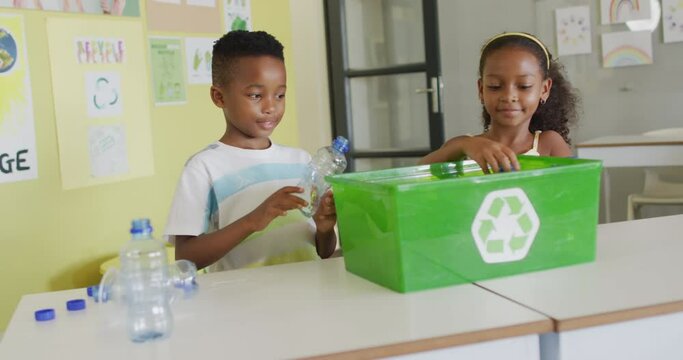 Video of happy african american girl and boy sorting plastic bottles for recycling in classroom