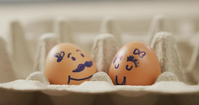 Video of close up of two brown eggs with drawn faces in egg carton background