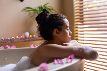 Side view of thoughtful biracial young woman looking through window blinds while leaning on bathtub