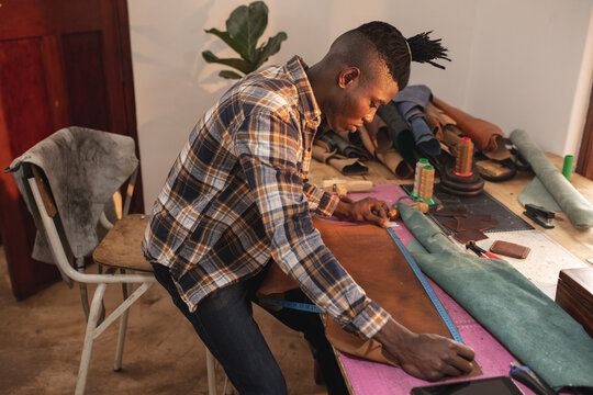 Side view of african american young craftsman measuring leather at workbench in workshop
