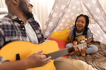 Cheerful african american daughter and father sitting with teddy bear and guitar in tent at home